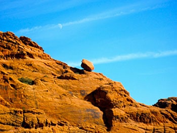 A boulder balances on the edge of a mountain as the daytime moon illuminates the sky.