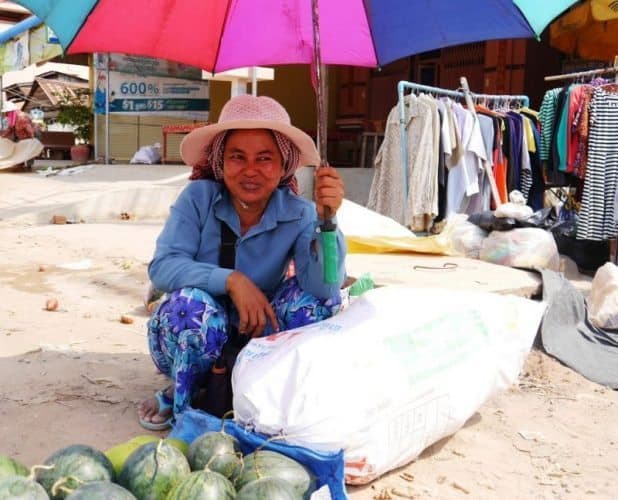 A street vendor shading herself from the harsh Cambodian sun while selling fresh produce and clothes