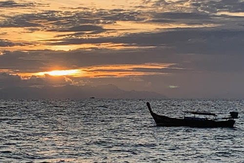 The golden sun peeks through at sunrise as a lone longtail sails by on Sunrise Beach.